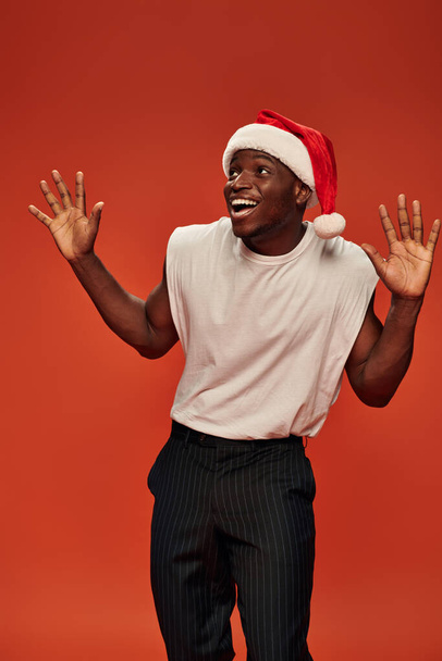 amazed and overjoyed african american man in santa hat showing wow gesture and looking away on red - Fotografie, Obrázek