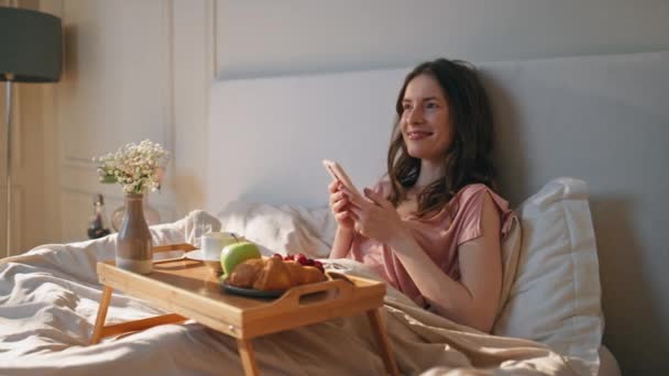 Vrolijk meisje dat thuis in bed ligt. Mooie romantische vrouw houdt mobiele telefoon chatten online. Ontspannen glimlachende vrouw liggend gezellige lakens genieten van het ontbijt. Zorgeloos model lachen look smartphone - Video