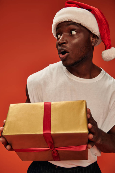 homme afro-américain étonné dans santa chapeau tenant boîte cadeau et détournant les yeux avec bouche ouverte sur rouge - Photo, image