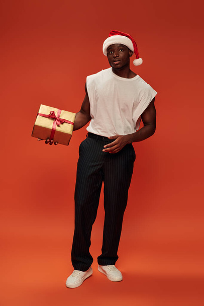 hombre afroamericano en ropa casual de moda y sombrero de santa con caja de regalo de oro en rojo, longitud completa - Foto, Imagen