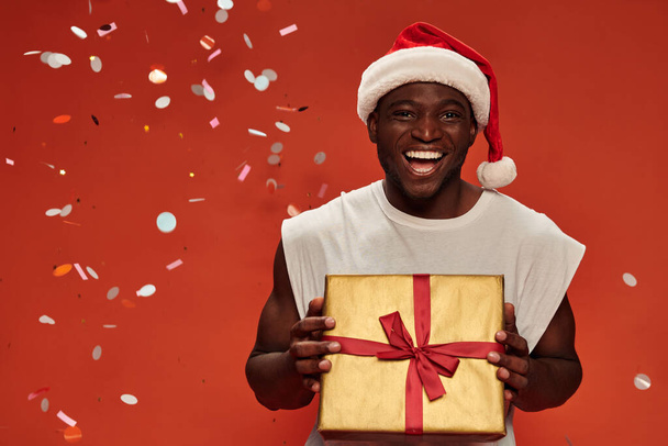 cheerful african american guy in christmas hat posing with gift boxes under falling confetti on red - Photo, Image