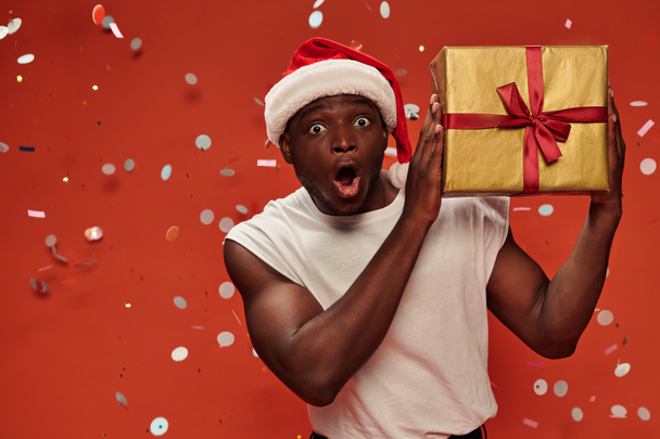 erstaunt afrikanisch-amerikanischer Mann in Weihnachtsmütze mit Geschenken und offenem Mund auf rotem Hintergrund mit Konfetti - Foto, Bild