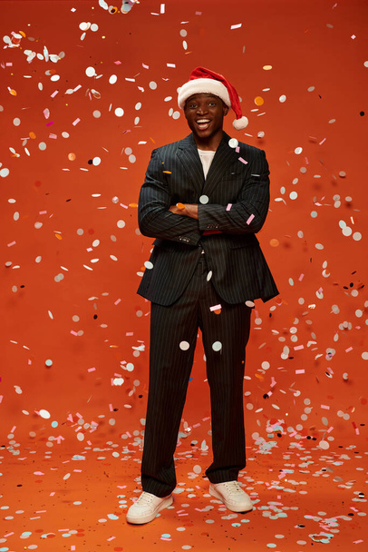 excited african american man in black suit and santa hat smiling under confetti on red backdrop - Foto, Imagem