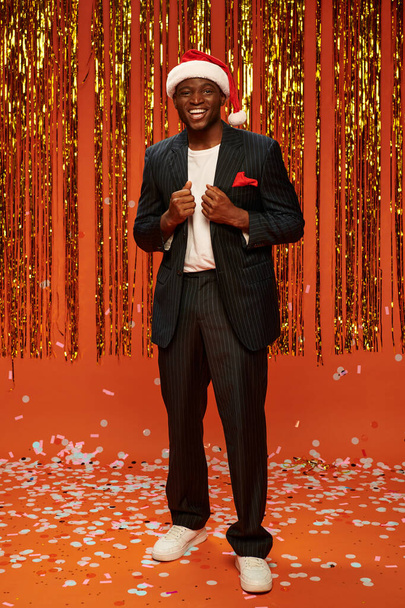 cheerful african american man in santa hat and black suit on orange backdrop with shiny decor - Fotoğraf, Görsel
