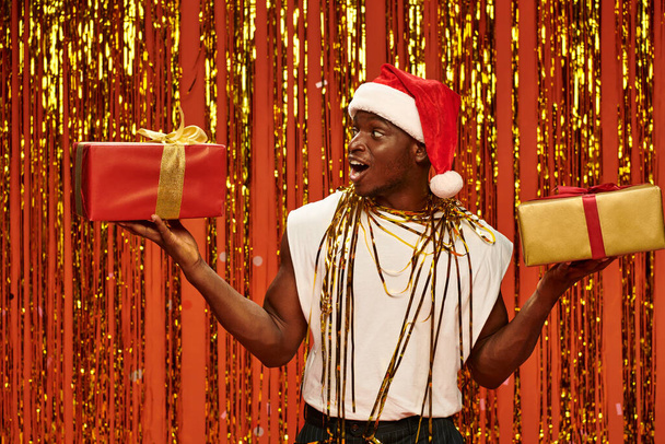 verbaasd afrikaanse amerikaanse man in santa hoed met kerst geschenken in de buurt glanzend tinsel op rode achtergrond - Foto, afbeelding