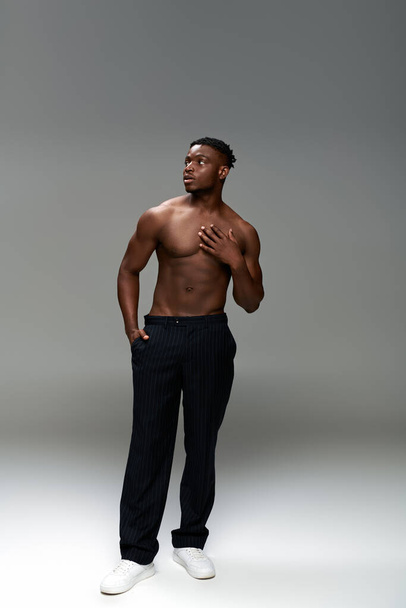 comprimento total de homem americano africano sem camisa e muscular com a mão em bolsos olhando para longe em cinza - Foto, Imagem