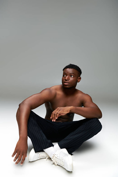 senza maglietta atletico afroamericano uomo in pantaloni neri seduto e guardando altrove su grigio, a tutta lunghezza - Foto, immagini