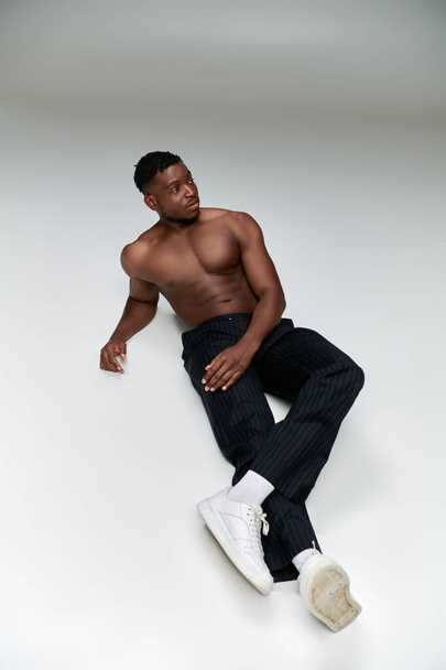 fiducioso modello di fitness africano americano senza maglietta in pantaloni neri seduto e guardando altrove sul grigio - Foto, immagini