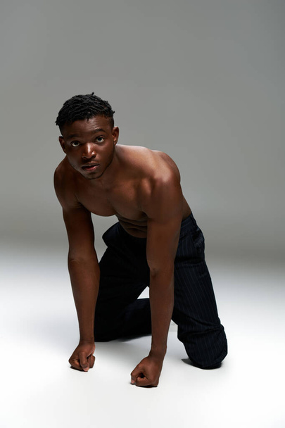 Мускулистый африканский американец в черных штанах, стоящий на коленях и смотрящий в камеру на сером - Фото, изображение