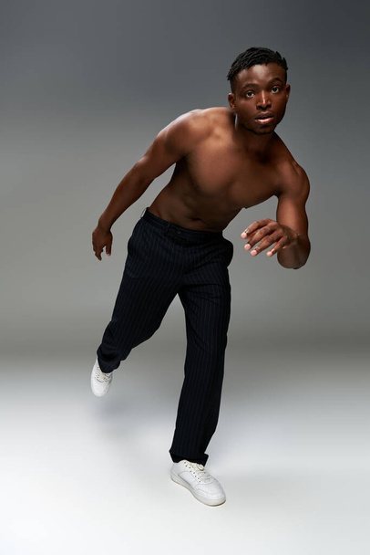 ημίγυμνος και αθλητικός Αφροαμερικανός άνδρας με μαύρο παντελόνι ποζάρει στο ένα πόδι σε γκρι φόντο - Φωτογραφία, εικόνα