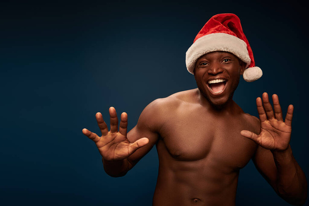overjoyed african american man with  muscular torso and santa hat gesturing on dark blue backdrop - 写真・画像