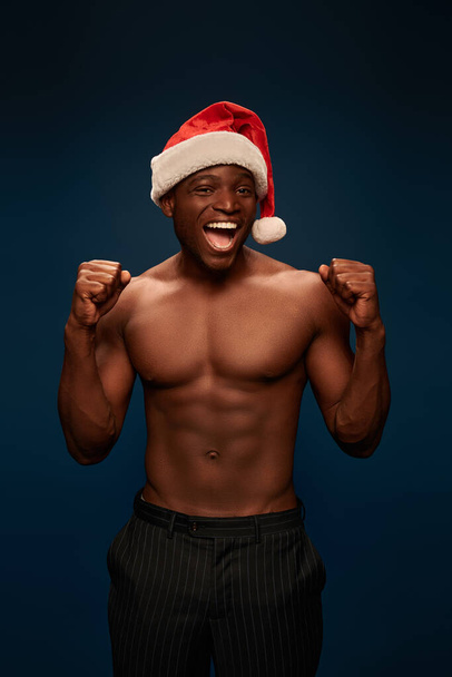 überglücklicher athletischer Afroamerikaner in Weihnachtsmannmütze mit Siegergeste vor marineblauem Hintergrund - Foto, Bild