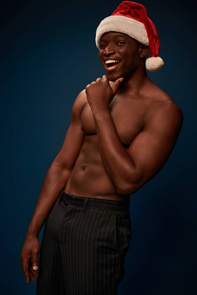 muscular and shirtless african american man in santa hat laughing at camera on navy blue backdrop - Фото, зображення