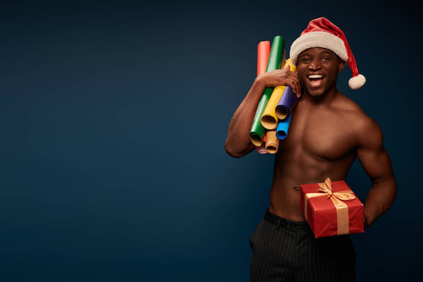 joyful and shirtless african american man in santa hat with wallpapers and gift box on dark blue - Foto, immagini