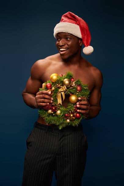 hombre afro americano sin camisa positivo en sombrero de santa con corona de Navidad mirando hacia otro lado en azul oscuro - Foto, Imagen