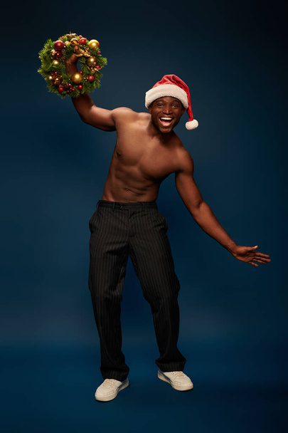 wesoły i bez koszuli Afrykański Amerykanin w kapeluszu Mikołaja pozowanie świąteczny wieniec na granatowym - Zdjęcie, obraz