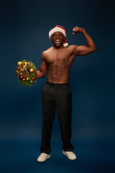 zdumiony i wysportowany Afrykański Amerykanin w kapeluszu Mikołaja pokazujący świąteczne wieńce na granatowym błękicie - Zdjęcie, obraz