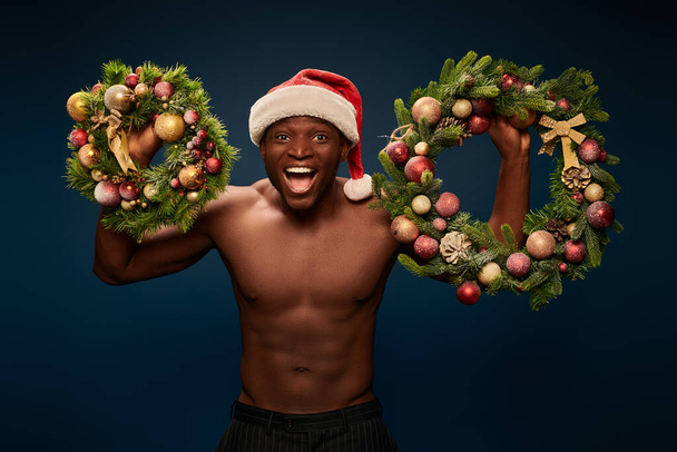 hemdlos energiegeladener afrikanisch-amerikanischer Mann mit Weihnachtsmütze, der Weihnachtskränze auf marineblauem Grund zeigt - Foto, Bild