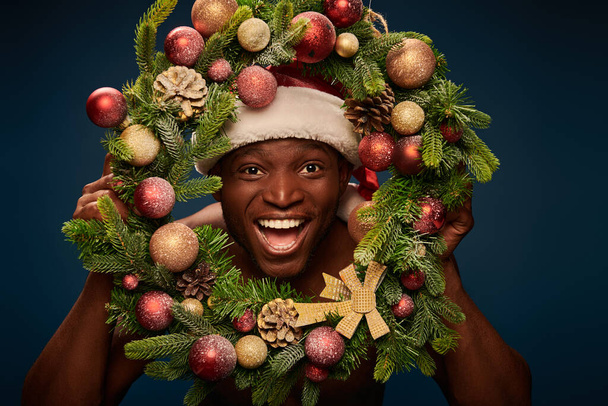サンタ帽子のアフリカ系アメリカ人男性が,暗い青色のクリスマスリースを通してカメラを見るのを楽しんだ. - 写真・画像