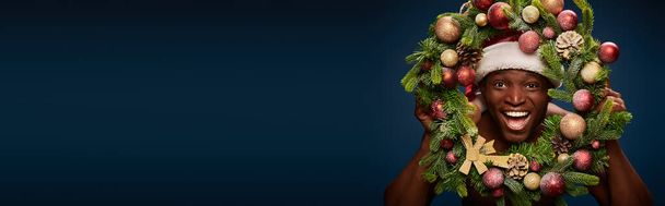 portrait of cheerful american man in santa hat and christmas wreath on dark blue backdrop, banner - Photo, Image