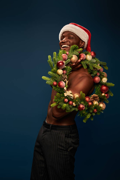 warm en gespierd Afrikaans Amerikaans model in santa hoed poseren met kerst krans op marine blauw - Foto, afbeelding