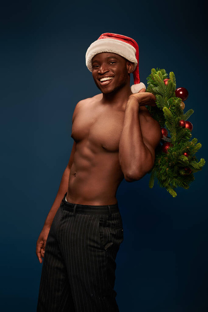 modelo afroamericano deportivo sin camisa en santa cap posando con corona de Navidad en azul oscuro - Foto, imagen