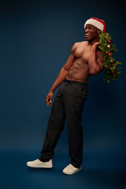 happy athletic african american man in santa hat holding christmas wreath on dark blue backdrop - 写真・画像