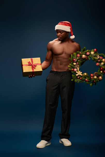 uomo afroamericano senza maglietta in cappello di Babbo Natale contenente scatola regalo e ghirlanda di Natale su blu scuro - Foto, immagini