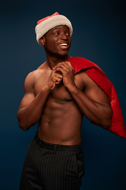 cheerful african american man with shirtless torso and christmas bag looking away on navy blue - Photo, Image