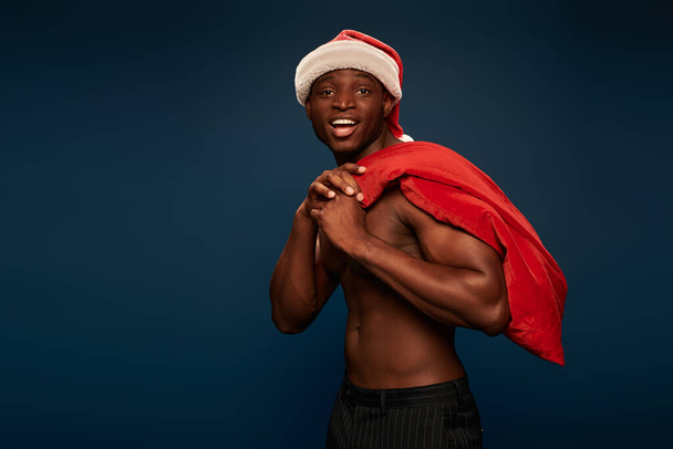 aufgeregter und starker afrikanisch-amerikanischer Mann mit Weihnachtsmütze, der eine Weihnachtstasche in der Hand hält und auf dunkelblau lächelt - Foto, Bild