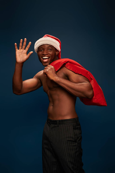 szczęśliwy i bez koszuli Afrykański amerykański człowiek z torbą świąteczną macha ręką na ciemnoniebieskim tle - Zdjęcie, obraz