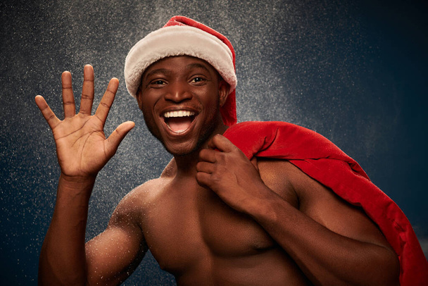 cheerful shirtless african american man with santa bag waving hand on dark blue snowy  backdrop - Foto, imagen