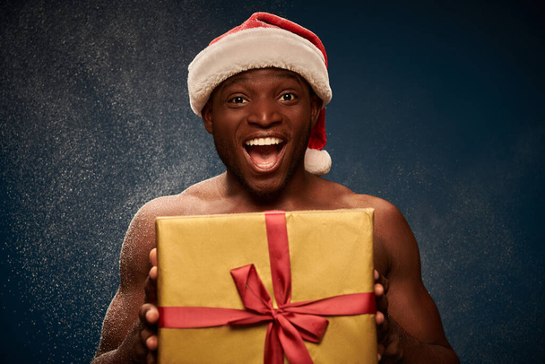 ενθουσιασμένος αθλητικός Αφροαμερικανός άνδρας σε καπέλο santa με γυαλιστερό κουτί δώρου σε σκούρο μπλε χιονισμένο φόντο - Φωτογραφία, εικόνα