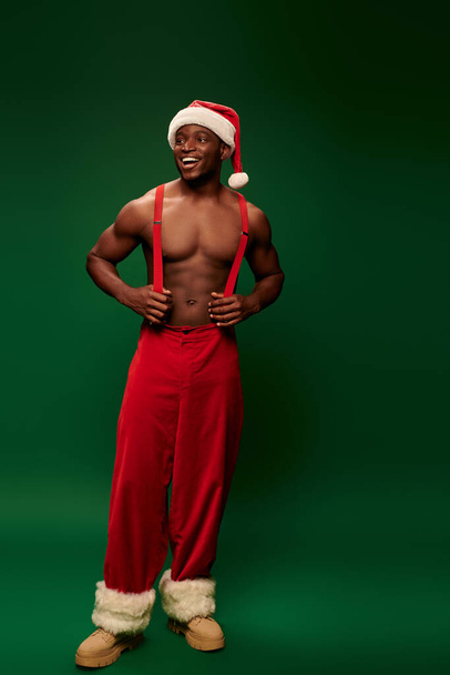 cheerful muscular african american man in santa hat and red pants with suspenders on green backdrop - Valokuva, kuva