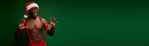 vidám sportos afro-amerikai férfi pózol télapó kalap és nadrág nadrágtartó zöld, banner - Fotó, kép