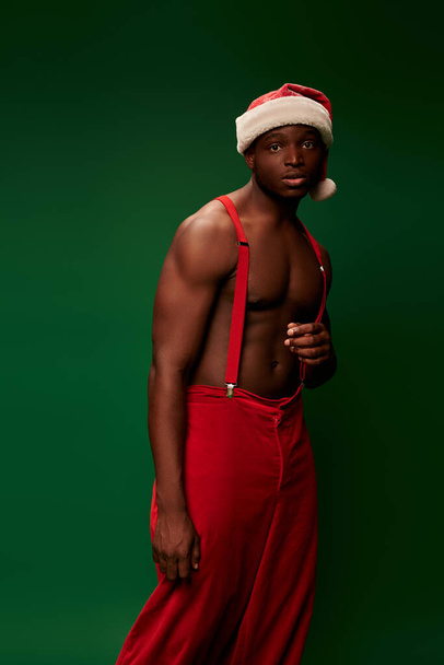 σκεπτικός Αφροαμερικάνος με γυμνό μυϊκό κορμό σε μαύρο καπέλο και κόκκινο παντελόνι σε πράσινο - Φωτογραφία, εικόνα