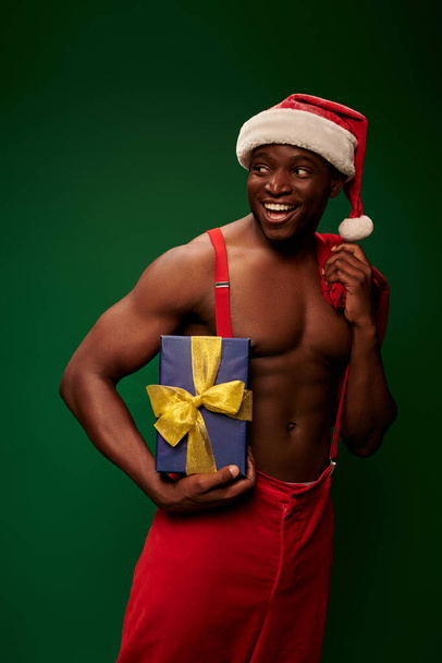cheerful athletic african american man in santa hat holding present and christmas bag on green - Photo, image