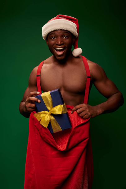 emocionado atlético afroamericano hombre en sombrero de santa con bolsa de Navidad y caja de regalo sobre fondo verde - Foto, Imagen