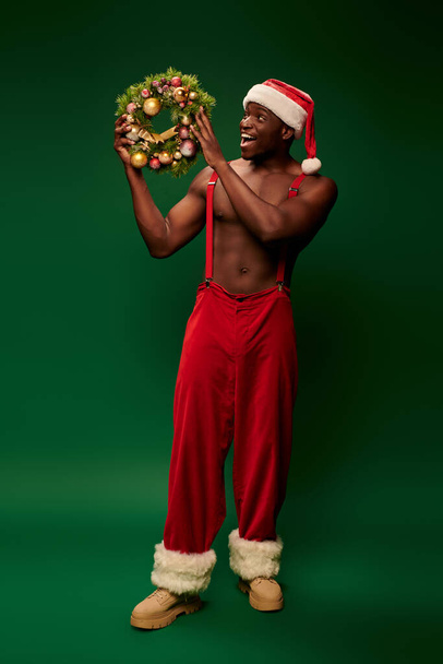 erstaunt afrikanisch-amerikanischer Mann mit Weihnachtsmütze und roter Hose schaut auf glänzenden Weihnachtskranz auf grünem Grund - Foto, Bild