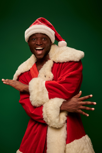 overjoyed african american man in santa claus costume gesturing and looking at camera on green - Photo, Image