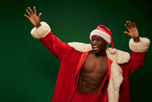 radosny african amerykański facet w kostiumie Bożego Narodzenia na ciało bez koszuli macha rękami na zielonym tle - Zdjęcie, obraz