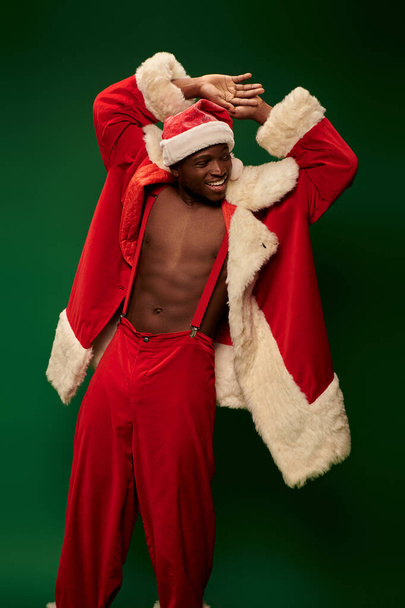 гарячий афроамериканський хлопець у різдвяному костюмі на без сорочки, посміхаючись і дивлячись на зелений - Фото, зображення