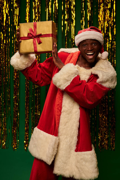 aufgeregter afrikanisch-amerikanischer Mann im Weihnachtskostüm zeigt Geschenk auf grünem und glänzendem Hintergrund - Foto, Bild