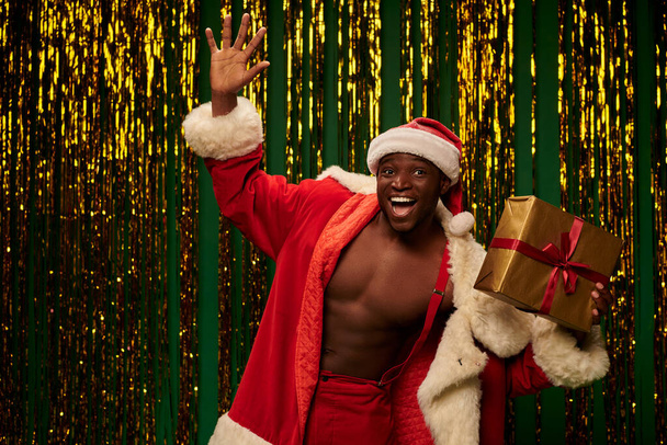 jolly african american man in christmas costume holding present and waving hand on shiny backdrop - Фото, изображение