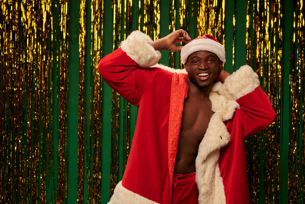 cheerful african american guy posing in santa costume on shirtless body near shiny tinsel on green - Foto, afbeelding