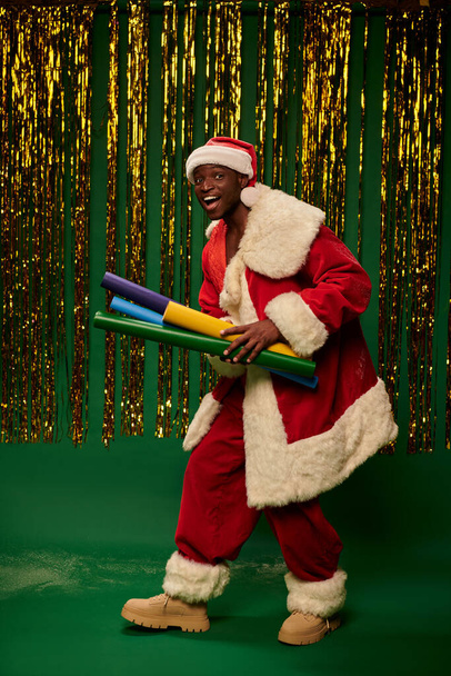 Noel Baba kostümlü neşeli Afro-Amerikalı adam. Renkli kağıtlarla süslenmiş yeşil arka planda süslemeleri var. - Fotoğraf, Görsel