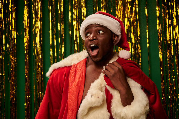 здивований афроамериканський чоловік у костюмі Санти з відкритим ротом та різдвяною сумкою біля золотої мішури - Фото, зображення