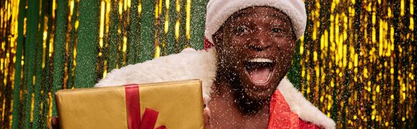 erstaunt afrikanisch-amerikanischer Mann im Weihnachtsmannkostüm mit Geschenkbox im schneebedeckten und glänzenden Atelier, Banner - Foto, Bild