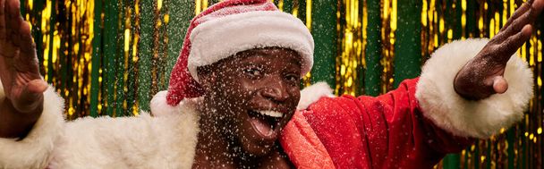 黄金の背景に雪の下でジェスチャーサンタ衣装の陽気なアフリカ系アメリカ人男性,バナー - 写真・画像