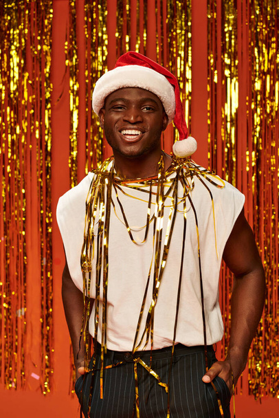 Noel Baba şapkalı ve beyaz kolsuz bluzlu neşeli Afro-Amerikalı adam kırmızı arka planda altın rengi süslemeler yapıyor. - Fotoğraf, Görsel